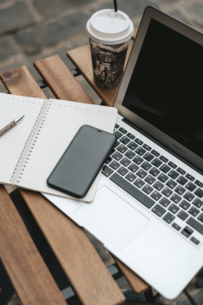From above of modern netbook placed on wooden table with opened notebook and cellphone near cup of hot coffee on terrace of cafeteria